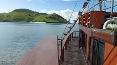 Labuan Bajo 'daki ahşap bir geminin sakin sulara bakan güvertesinden manzara ve açık mavi gökyüzünün altında yemyeşil bir tepe..