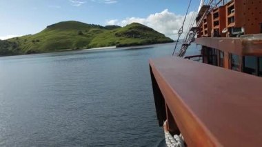 Labuan Bajo 'daki bir geminin güvertesinden manzaralı, sakin suları ve arkaplanda parçalı bulutlu bir gökyüzünün altında yemyeşil bir adayı gösteriyor..