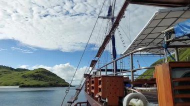 Güverteyi ve direği gösteren bir teknenin manzarası yeşil tepelerin ve parçalı bulutlu gökyüzünün zeminine karşı. Sakin su bulutları yansıtır, sakin bir atmosfer yaratır..