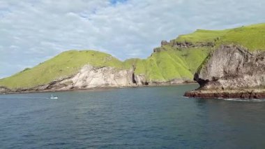 Issız bir ada. Bereketli yeşil tepeler ve kayalık kayalıklardan oluşan kıyı manzarası. Üzerinde parçalı bulutlu bir gökyüzü bulunan sakin mavi sularda yol alan küçük bir tekne görülüyor..