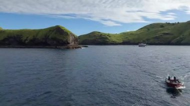 Issız Ada - sakin mavi bir körfezi çevreleyen yemyeşil tepelerin olduğu sakin bir sahil manzarası. Küçük bir tekne denizde yüzüyor, daha büyük bir gemi de uzak bir yere demir atmış..