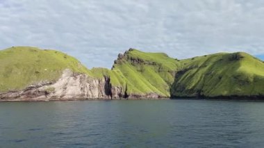 Issız Ada - Sakin bir denizden keskin bir şekilde yükselen yemyeşil tepelerin manzarası, tabanında kayalık kayalıklar görülüyor. Gökyüzü parçalı bulutlu, huzurlu ve resimsel bir manzara yaratıyor..