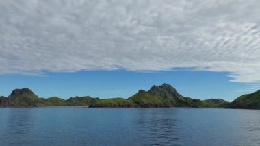 Parçalı bulutlu bir gökyüzünün altında yemyeşil dağlarla çevrili sakin bir su kütlesinin olduğu sakin bir manzara. Sahne, doğanın güzelliğini canlı renklerle ve huzurlu bir atmosferle yakalıyor..