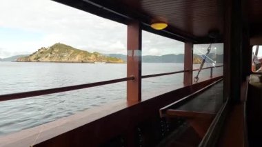 Teknenin içinden manzaralı bir manzara, sakin bir su kütlesi ve tepelerle çevrili uzak bir adayı gösteriyor. Teknenin iç kısmında ahşap paneller ve geniş pencereler var..