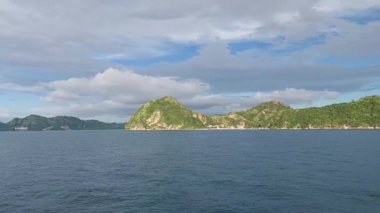 Bereketli yeşil tepeler ve parçalı bulutlu gökyüzü ile sakin bir denizin manzarası. Su, bulutları ve tepelerin yeşilliğini yansıtır. Huzurlu bir atmosfer yaratır..