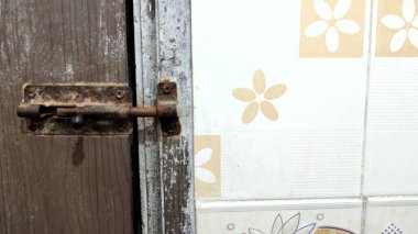 A close-up view of a rusty door latch on a wooden door, with a tiled wall featuring floral patterns in the background. The latch shows signs of wear and age, contrasting with the clean tiles. clipart