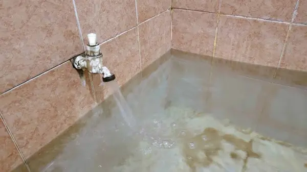 stock image A close-up view of a water tap in a tiled bathroom, with water flowing out and pooling on the floor. The tiles are light brown, and the water is slightly murky, reflecting the surroundings.