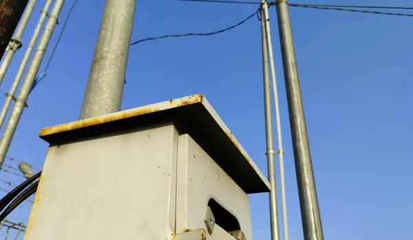 Arka planda açık mavi gökyüzü olan bir direğe monte edilmiş yıpranmış bir elektrik kutusunun yakın çekim görüntüsü. Kutuda pas ve aşınma izleri var, metal direkler ve tellerle çevrili..
