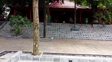 Sragen, Endonezya: Ağaçlarla çevrili kırmızı çatılı geleneksel bir ahşap bina. Yapıda, yakınındaki taş duvarlar ve yeşil çöp kutularıyla ona doğru giden basamaklar yer alıyor. İnsanlar basamaklara oturmuş, huzurlu ortamın tadını çıkarıyorlar..