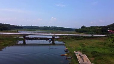 Köprü üzerinden geçen bir nehrin manzarası. Suyun etrafını saran yemyeşil tarlalar ve ağaçlar, üzerinde açık mavi bir gökyüzü var. Köprüde birkaç araç görünüyor ve küçük bir tekne nehir kıyısına demirlemiş durumda..