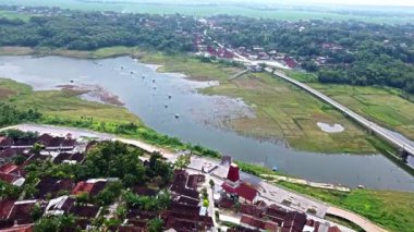 Bir nehir, yeşil tarlalar ve küçük bir köyden oluşan sakin bir manzaranın havadan görünüşü. Nehir, yemyeşil bitki örtüsü ve tarım arazisiyle çevrili olarak olay yerinden akar. Nehir kıyısı boyunca evler görülebiliyor. Bölgede bir yol dönüyor.