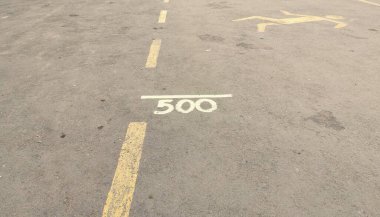 Sarı bir çizgi ve 500 'ün beyaza boyandığı bir otopark yüzeyi yakın çekim görüntüsü. Asfalt yıpranmış ve bazı kirli yamalar var, diğer park alanlarının belirsiz çizgileri görünür.