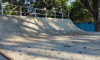 Ağaçlar ve yapraklarla çevrili bir parkta beton bir kaykay rampası. Rampa boş, yüzeye saçılmış yapraklar ve güneş ışığı gölgeler bırakıyor..