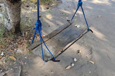 Kırsal ahşap bir salıncak, mavi iplerden sarkan, dağınık yapraklı toprak bir patikaya kurulmuş. Salıncak basit ve yıpranmış. Nostaljik bir açık hava ortamı..