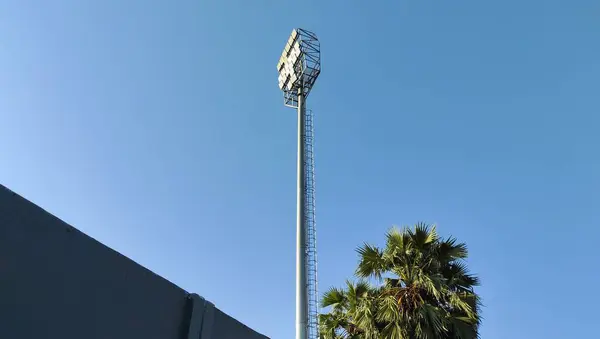 Önünde palmiye ağacı olan açık mavi gökyüzüne karşı yüksek bir stadyum ışık kulesi..