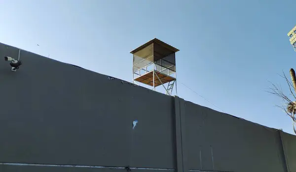 Açık mavi gökyüzüne karşı gri bir duvarda uzun bir gözetleme kulesi. Kulede ahşap bir platform ve duvarında güvenlik kamerası olan bir çatı var..