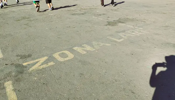 Asfalt zeminde yürüyen insanlarla dolu kalabalık bir alan. Zemin 'ZONA LARI' (çalışma bölgesi) yazan sarı işaretlerle solmuş. Kamera tutan bir kişinin gölgesi görünür..