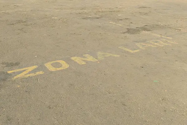 'ZONA LARI' (çalışma bölgesi) yazılı soluk sarı boya ile beton bir yüzeye yakından bakmak. Toprak tozlu ve yıpranmış görünüyor. Bu da yoğun yaya trafiğini gösteriyor..