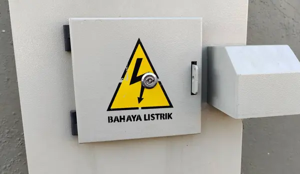 İçinde sarı üçgen ve şimşek işareti olan gri bir elektrik kutusu. Tabelada 'BAHAYA LISTRIK' yazıyor, elektrikten kaynaklanan tehlikeyi gösteriyor. Kutu duvara monte edilmiş..
