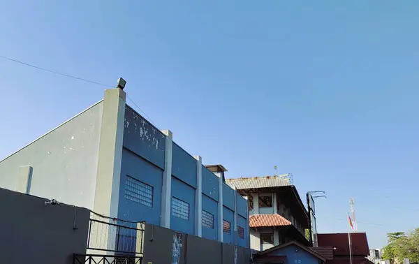 Düz çatılı gri bir binanın üzerinde açık mavi bir gökyüzü ve eğimli bir çatısı olan komşu bir yapı. Sahne, şehir mimarisini minimum bitki örtüsüyle yakalıyor..
