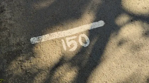 Çevredeki ağaçların gölgeleriyle kısmen gizlenmiş, toprak bir yolda '150' numaralı beyaz boyanın yakın plan görüntüsü..