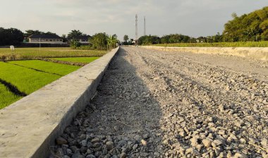 Yeşil pirinç tarlalarının yanında inşaat halindeki çakıllı bir yol. Sahnede açık bir gökyüzü ve dokulu çakıl yüzeyine odaklı uzak telekomünikasyon kuleleri yer alıyor..