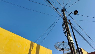 Birden fazla elektrik hattının bulunduğu açık mavi bir gökyüzü. Ön planda sarı bir duvar var, ve yakınlarda bir uydu anteni var, şehir manzarası yaratıyor..