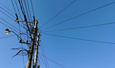 Açık mavi gökyüzü boyunca uzanan sayısız elektrik hattı olan uzun bir elektrik direği. Direk bir sokak lambası ve çeşitli kablolar ile donatılmıştır, gökyüzünün arka planına karşı karmaşık bir ağ oluşturur.