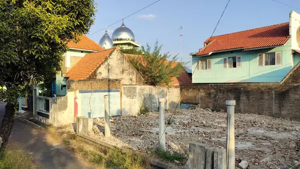 Etrafı konut binalarıyla çevrili boş bir arazi. Arka planda, muhtemelen bir caminin parçası olan iki kubbeli yapı ve yukarıda açık mavi bir gökyüzü var..