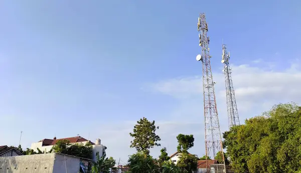 stock image Two tall telecommunications towers against a clear blue sky, surrounded by trees and buildings. The scene captures a mix of urban and natural elements.