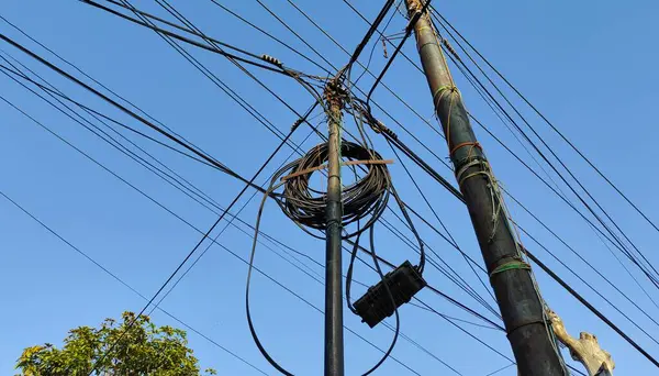 Açık mavi gökyüzüne karşı birbirine dolanmış elektrik kablolarıyla elektrik direkleri manzarası. Görüntü, kentsel altyapının karmaşıklığını yakalıyor, güç kablolarını ve kablolarını gösteriyor.
