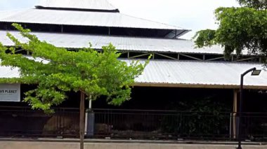 Ayırt edici piramit şekilli çatısı olan, metalik gri çatı panelleri olan büyük bir binanın havadan görünüşü. Çevresi yemyeşil ve ağaçlarla dolu, bulutlu bir gökyüzünün altında..