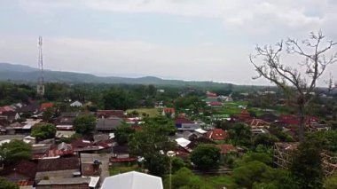 Geleneksel evleri, yeşil tarlaları ve parçalı bulutlu gökyüzünün altındaki uzak dağları olan küçük bir kasabanın panoramik manzarası..