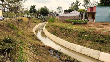 Yeşil bir arazide uzanan dar, beton kaplı bir drenaj kanalı. Bir tarafta ağaçlar ve çalılar, diğer tarafta bahçeli evler var. Gökyüzü parçalı bulutlu.