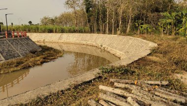Ağaçlar ve bitki örtüsüyle çevrili taş duvarlarla kaplı kıvrımlı bir su kanalı. Su sakin ve çevredeki yeşilliği yansıtıyor. Günlükler yakınlarda istiflenmiş, son temizliği ya da günlüğü gösteriyor.