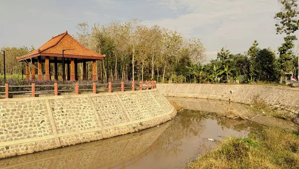 Yeşillik ve nehirle çevrili geleneksel bir taraçanın yer aldığı sakin bir park sahnesi..