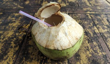 İçinde pipet olan taze bir hindistan cevizi, kırsal ahşap bir yüzeyde duruyor. Hindistan cevizi kısmen açılır ve içindeki kahverengi sıvı ortaya çıkar. Hindistan cevizi kabuğunun dokusu pürüzsüz samanla çelişen pürüzsüz ve doğal..