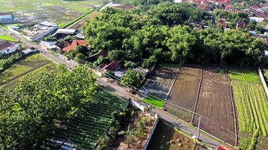 Tarlaların, evlerin ve yeşilliklerin yer aldığı kırsal bir arazinin havadan görünüşü. Sahnede ekili arazi, bir yol ve yerleşim ve tarım alanlarının karışımı yer alıyor..