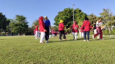 Pati, Endonezya - 08092024: Güneşli bir parkta açık hava aktivitelerine katılan kırmızı ve mavi giysili bir grup insan. Bazı bireyler bir oyuna dahil olur..