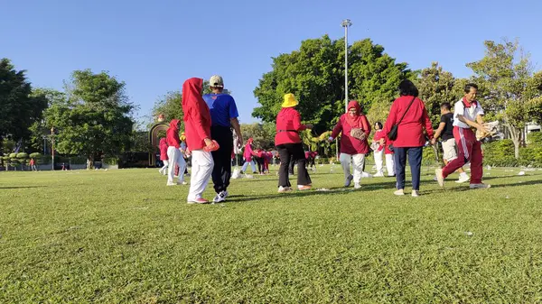 Pati, Endonezya - 08092024: Güneşli bir parkta açık hava aktivitelerine katılan kırmızı ve mavi giysili bir grup insan. Bazı bireyler bir oyuna dahil olur..