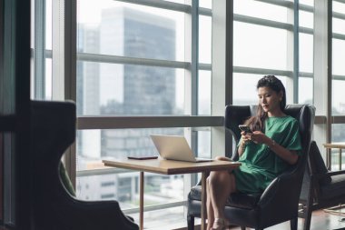 Ofis salonunda telefon ve laptop kullanan Asyalı bir kadın.