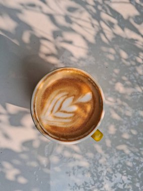 Sarı bardakta sıcak kapuçino, güneş ışığına maruz kalmış, beyaz bir arka planda izole edilmiş.