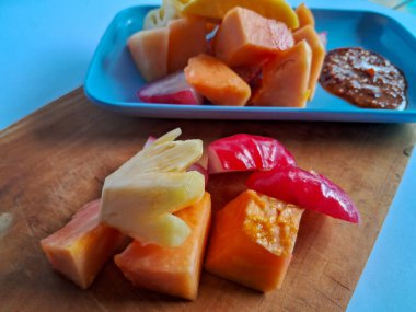 Bir parça mango, kavun, ananas, mavi bir tabakta papaya tuzlu, tatlı ve ekşi aromalı sosla birlikte beyaz tabaktaki meyve parçaları.
