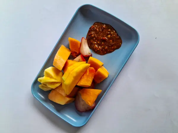 Bir parça mango, kavun, ananas, mavi bir tabakta papaya tuzlu, tatlı ve ekşi aromalı sosla birlikte beyaz tabaktaki meyve parçaları.