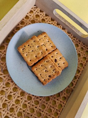 Minimalist mavi bir tabakta servis edilen, dokuma ahşap tepsiye konmuş, siyah susam tohumlu kare krakerler, konforlu ve sade bir yemek sunumu yaratır..