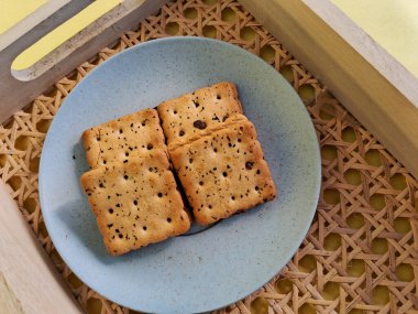 Minimalist mavi bir tabakta servis edilen, dokuma ahşap tepsiye konmuş, siyah susam tohumlu kare krakerler, konforlu ve sade bir yemek sunumu yaratır..