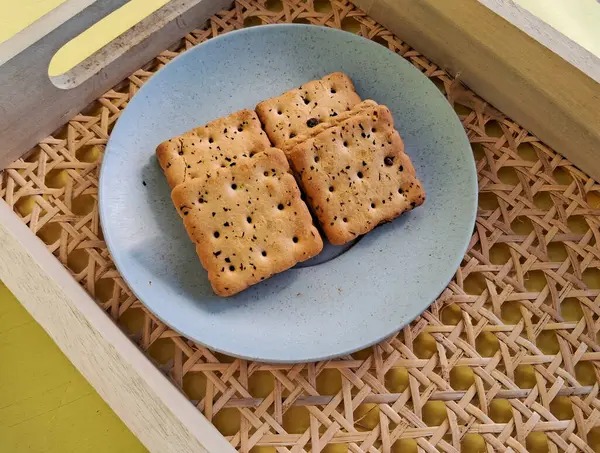 Minimalist mavi bir tabakta servis edilen, dokuma ahşap tepsiye konmuş, siyah susam tohumlu kare krakerler, konforlu ve sade bir yemek sunumu yaratır..