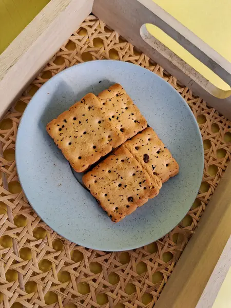 Minimalist mavi bir tabakta servis edilen, dokuma ahşap tepsiye konmuş, siyah susam tohumlu kare krakerler, konforlu ve sade bir yemek sunumu yaratır..