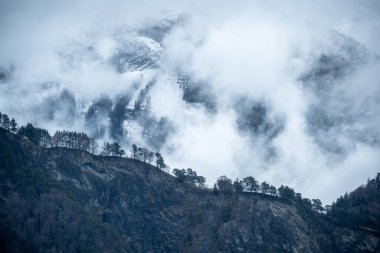 Siluetli ağaçlarla kaplı engebeli dağ sırtları sisli, dağınık bulutlar devasa bir dağ yamacını ortaya çıkarıyor. Koyu mavi tonlar ve yumuşak ışık gizemli ve karamsar atmosferi güçlendirir..