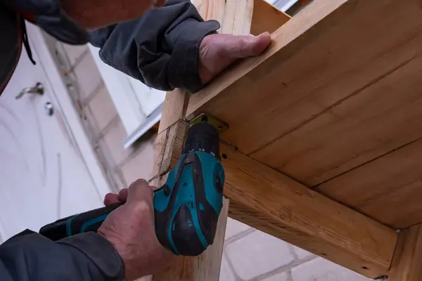 Bir adam matkapla çalışır. Usta elektrikli matkap kullanarak ahşap bir bank kuruyor..